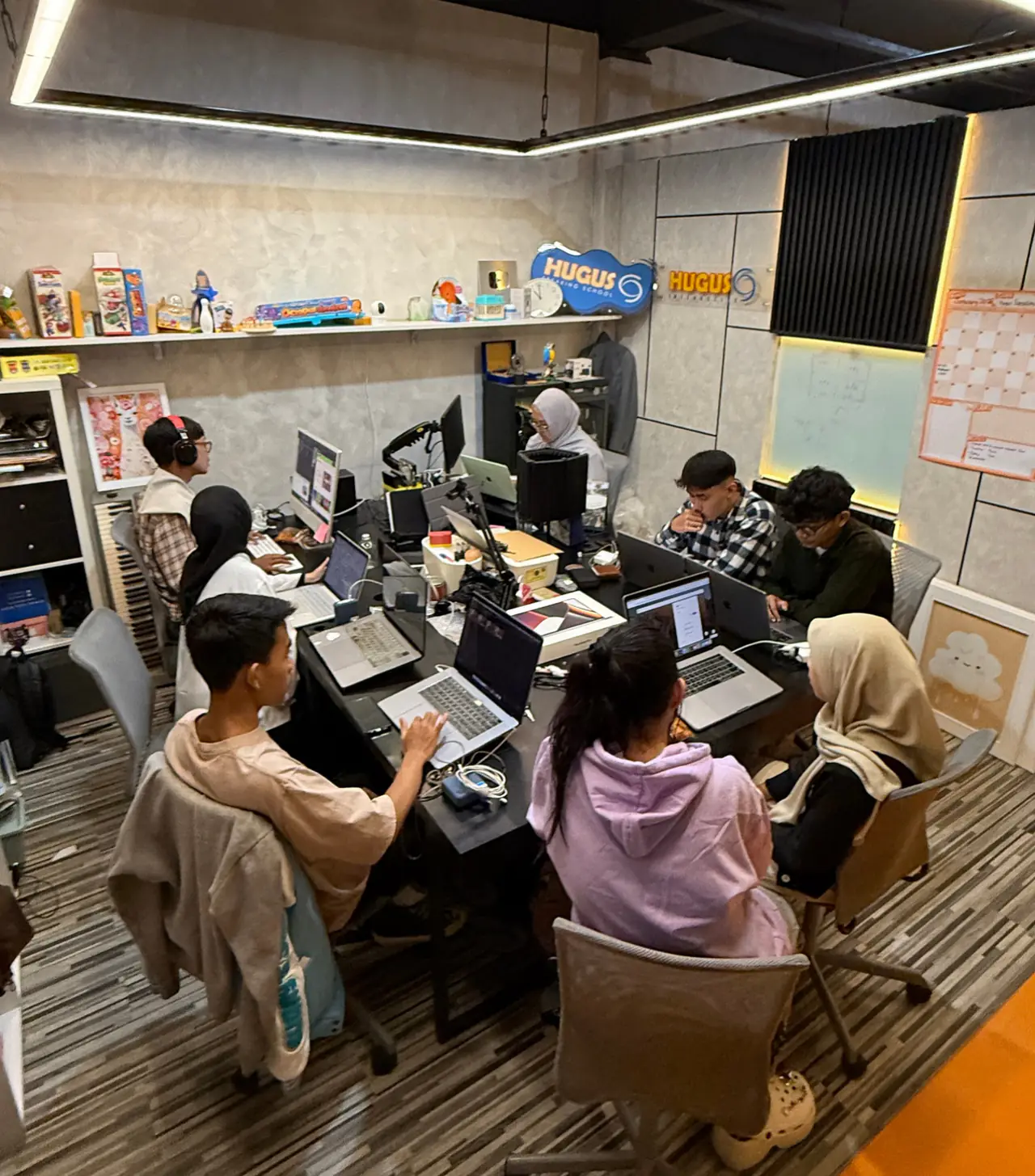 Tim muda bekerja dengan laptop di meja panjang studio HUGUS, dengan papan nama HUGUS terpajang di rak.