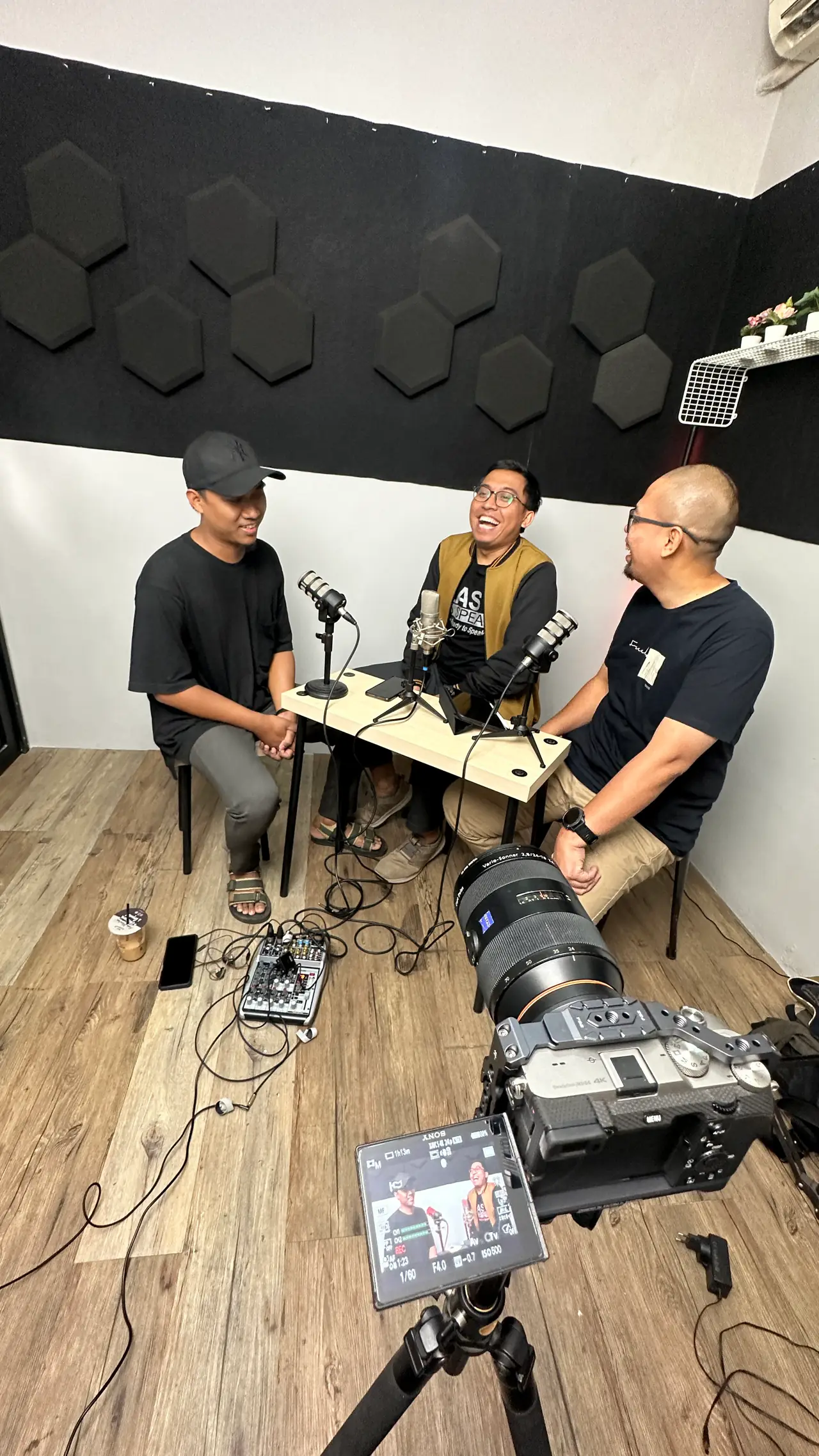 Rekaman podcast tiga orang di studio berpanel akustik, kamera bertripod di latar depan.