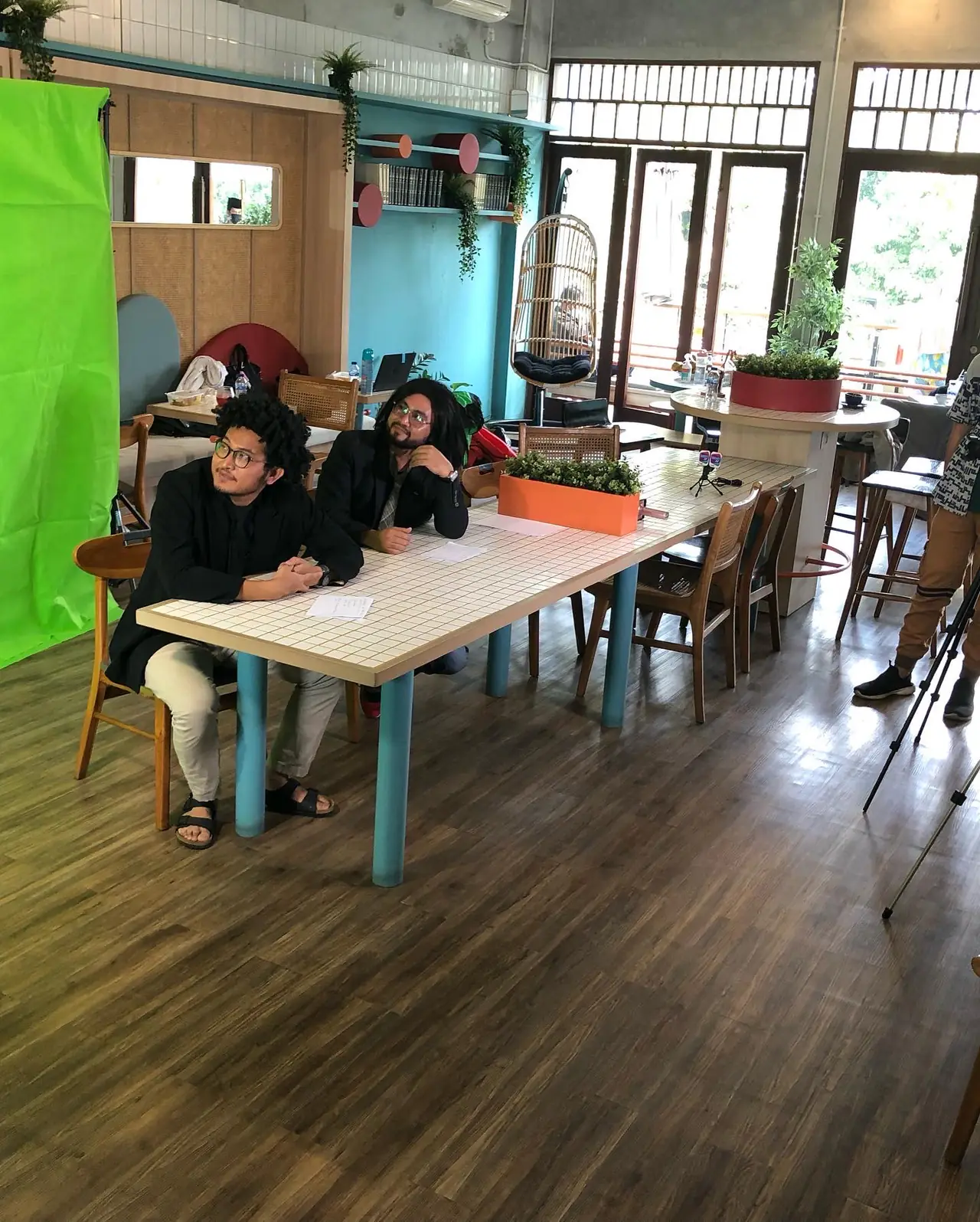 Dua orang duduk di meja panjang di ruang studio berlatar layar hijau.