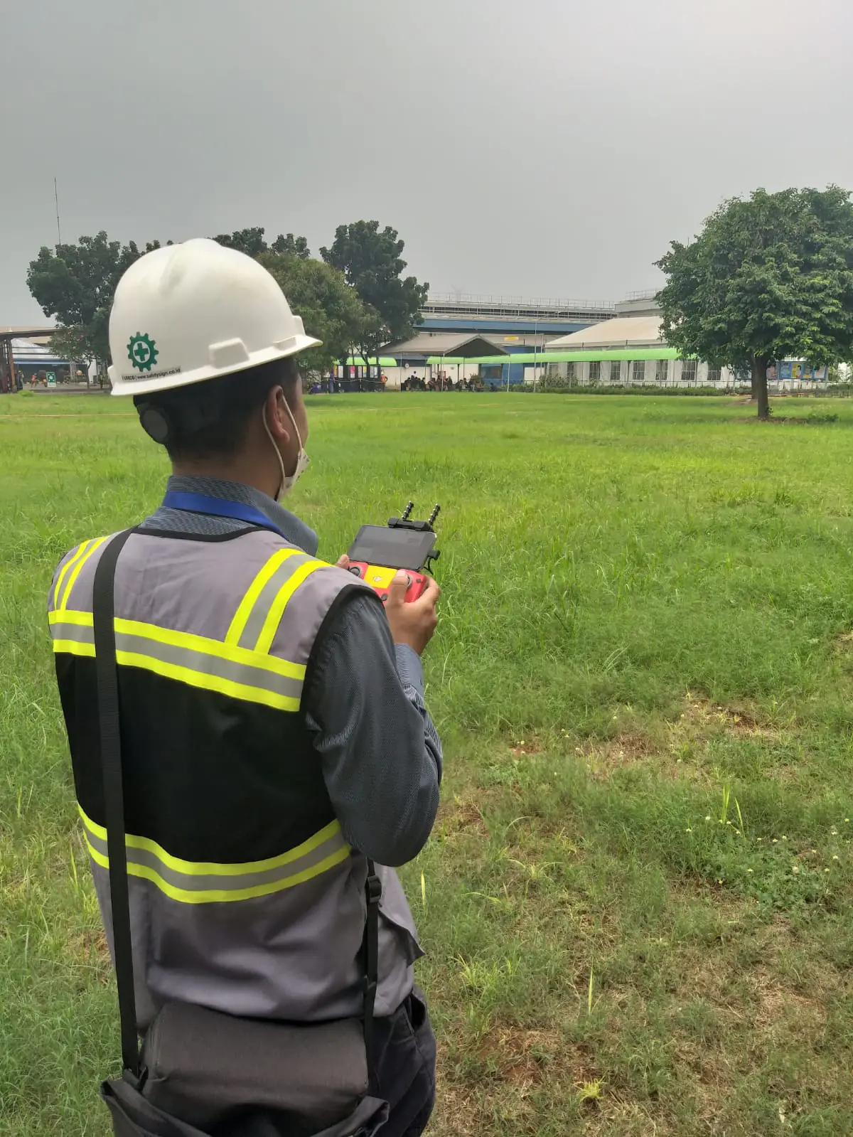 Anggota tim berhelm proyek dan berompi keselamatan memegang remote kendali di lapangan terbuka.