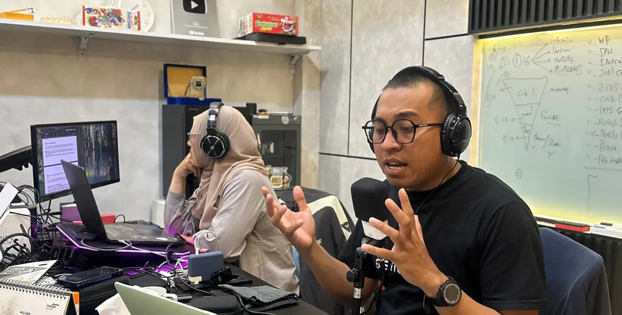 Pria yang memakai headphone berbicara di depan mikrofon dalam sesi rekaman, sementara rekan berhijab memantau layar komputer.