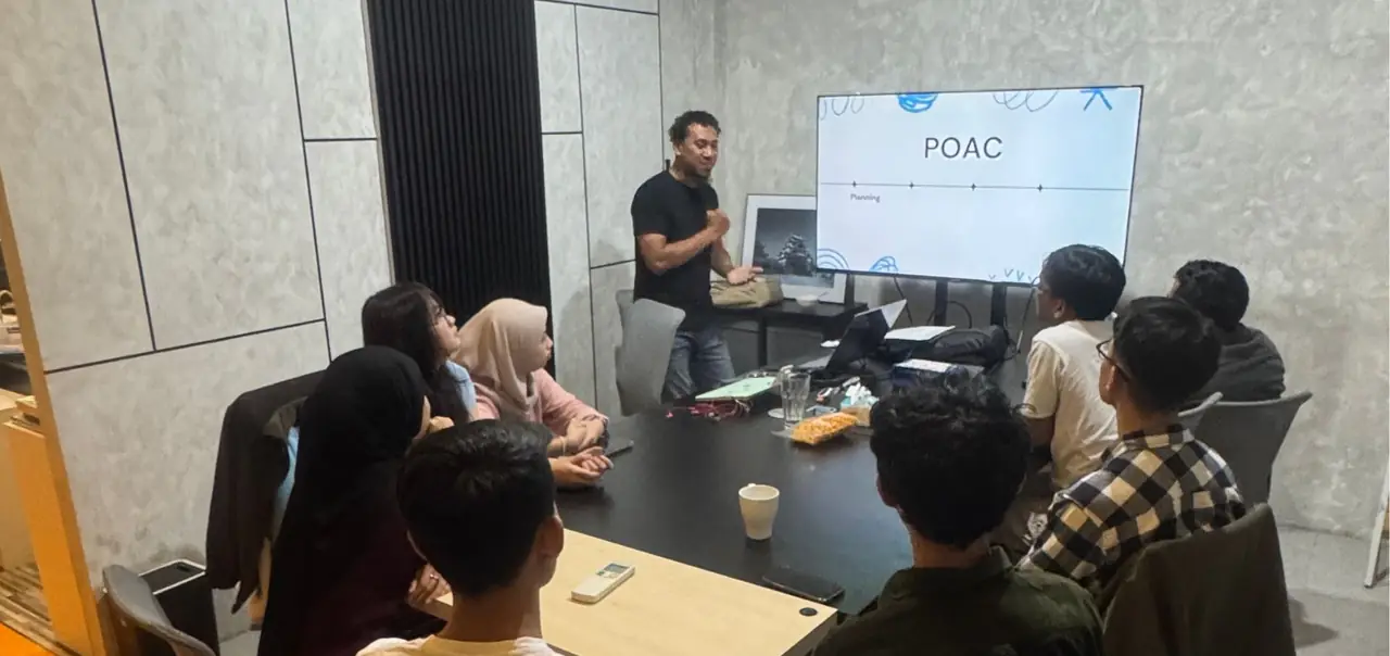 Pembicara berdiri di depan layar presentasi di ruang rapat, sementara peserta muda duduk mengelilingi meja.